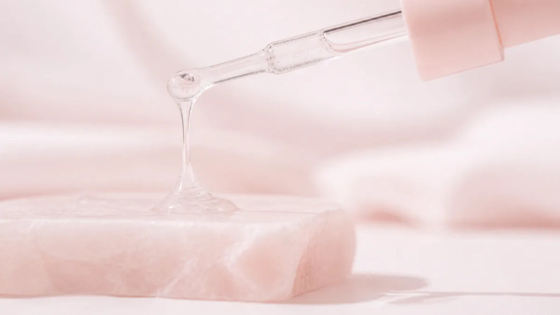 A clear viscous gel drawn into a slow thread between a glass dropper and a pale rose stone slab