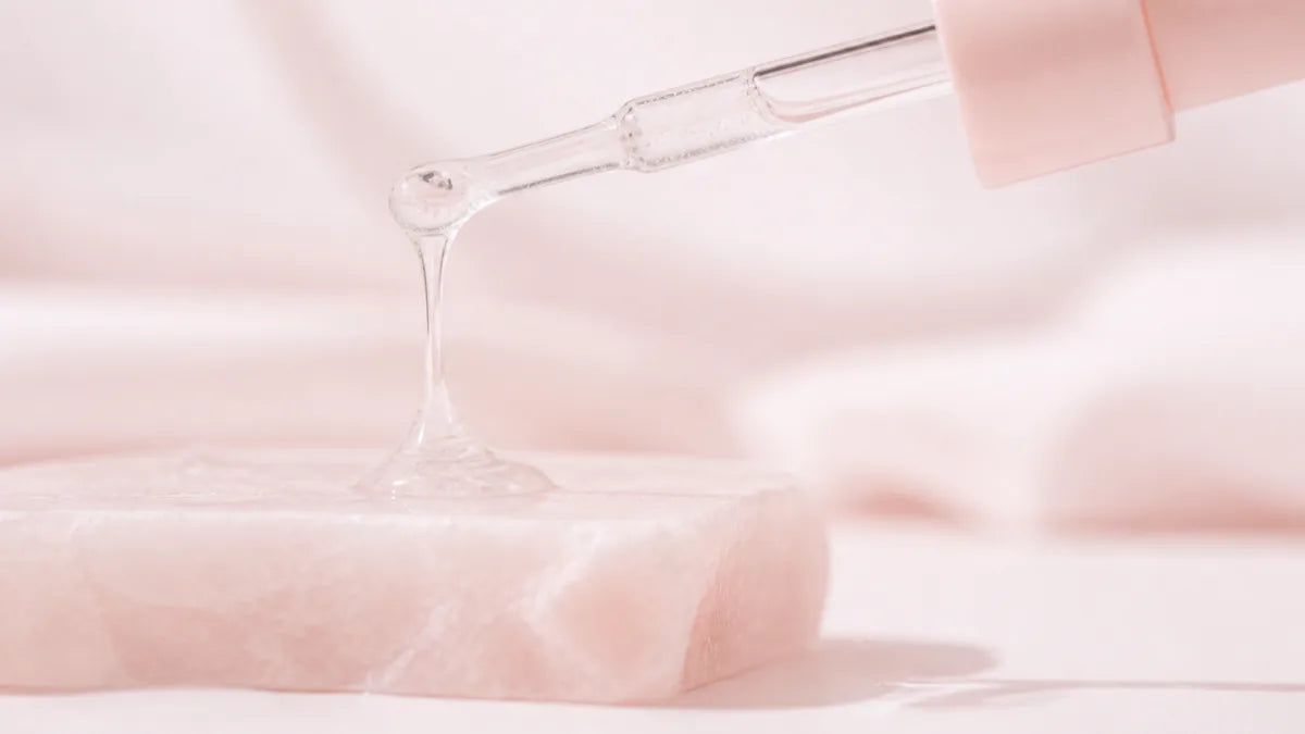 A clear viscous gel drawn into a slow thread between a glass dropper and a pale rose stone slab