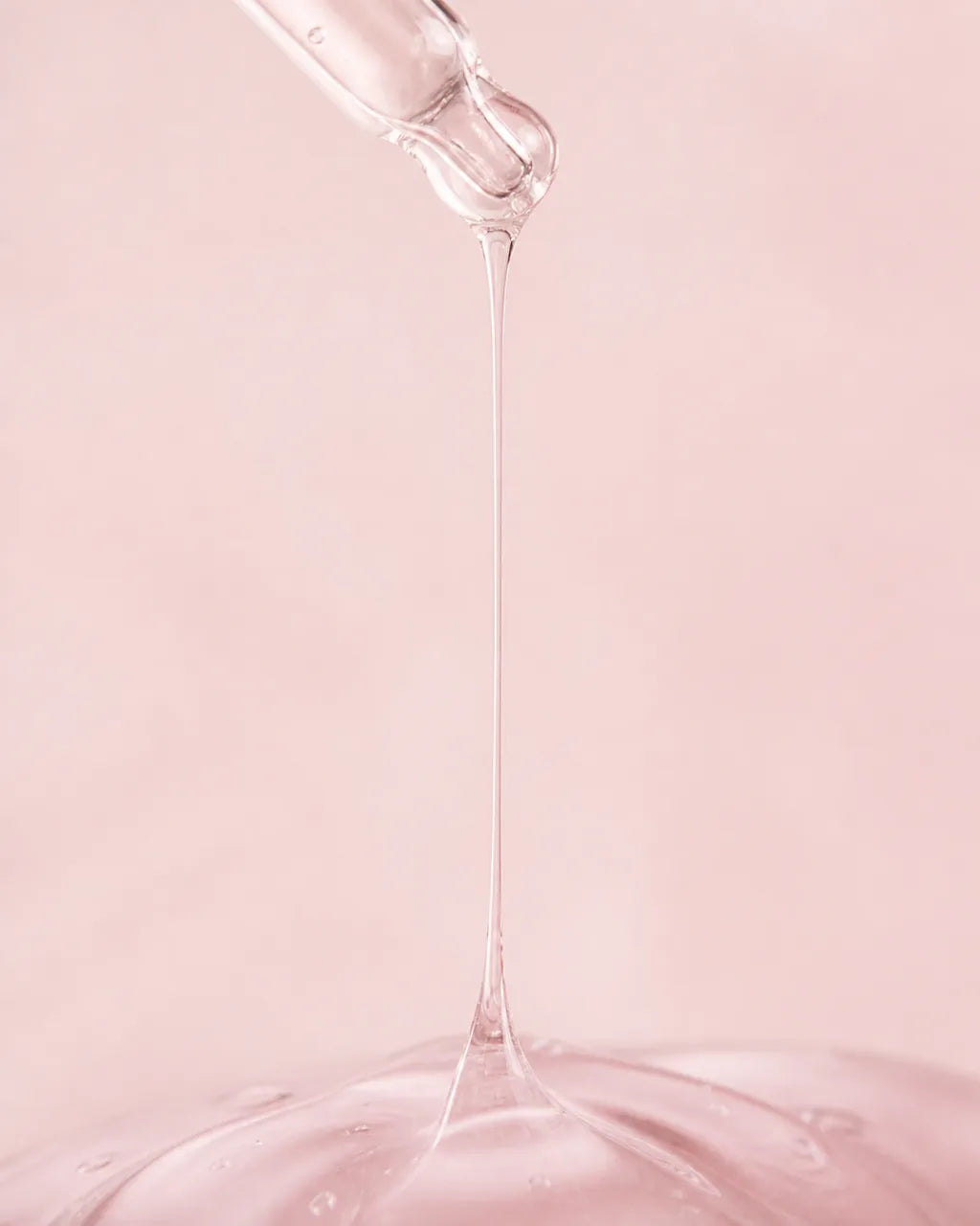 Close up of a clear viscous gel drawn into a single fine thread above a blush background